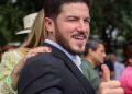 Man in a dark suit smiles and gives a thumbs-up at an outdoor gathering, with others visible in the blurred background.