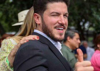 Man in a dark suit smiles and gives a thumbs-up at an outdoor gathering, with others visible in the blurred background.