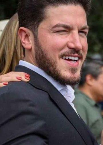 Man in a dark suit smiles and gives a thumbs-up at an outdoor gathering, with others visible in the blurred background.