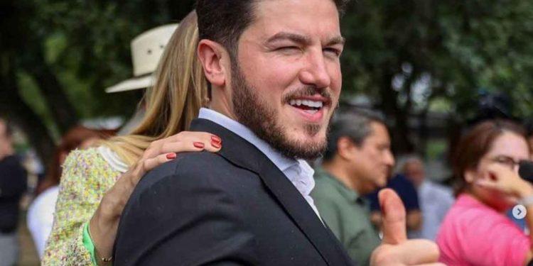 Man in a dark suit smiles and gives a thumbs-up at an outdoor gathering, with others visible in the blurred background.