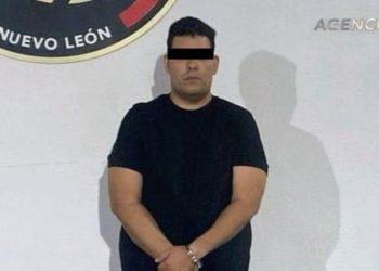 Man in a black T-shirt with hands cuffed in front, standing against a wall; eyes obscured with a black bar in what appears to be a police booking photo.