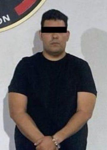 Man in a black T-shirt with hands cuffed in front, standing against a wall; eyes obscured with a black bar in what appears to be a police booking photo.