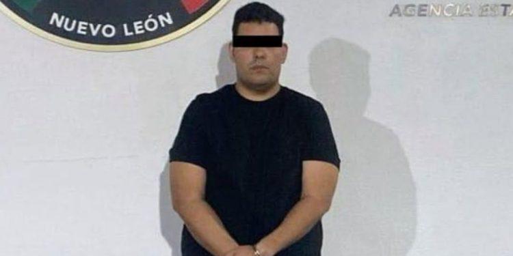 Man in a black T-shirt with hands cuffed in front, standing against a wall; eyes obscured with a black bar in what appears to be a police booking photo.