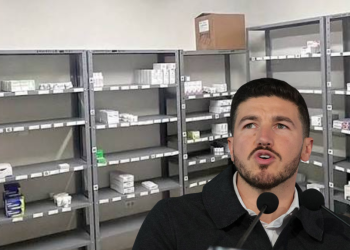 Man speaking into two microphones in a room with empty metal pharmacy shelves and scattered medicine boxes behind him