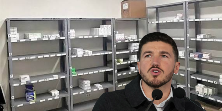 Man speaking into two microphones in a room with empty metal pharmacy shelves and scattered medicine boxes behind him