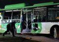 Nighttime scene of a green and white bus with shattered windows and a person crouching near the front door after a crash