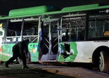 Nighttime scene of a green and white bus with shattered windows and a person crouching near the front door after a crash