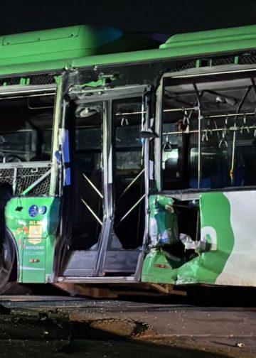 Nighttime scene of a green and white bus with shattered windows and a person crouching near the front door after a crash