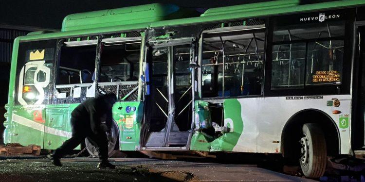 Nighttime scene of a green and white bus with shattered windows and a person crouching near the front door after a crash